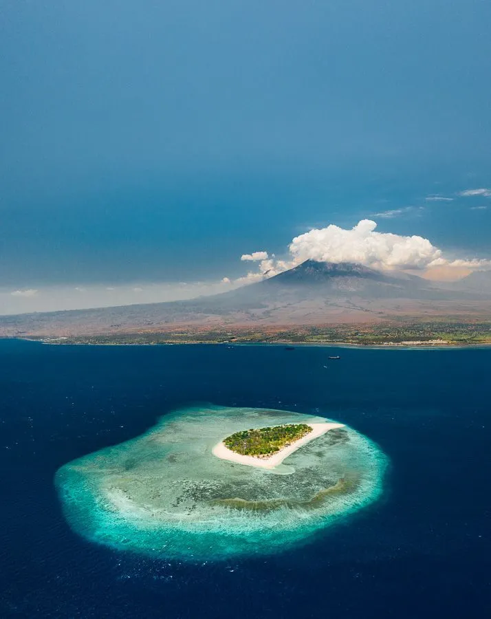 Pulau kecil berpasir putih di laut biru dengan gunung di kejauhan